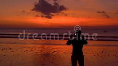 海滩上一个男人在美丽的日落时用<strong>手机</strong>拍照的<strong>剪影</strong>